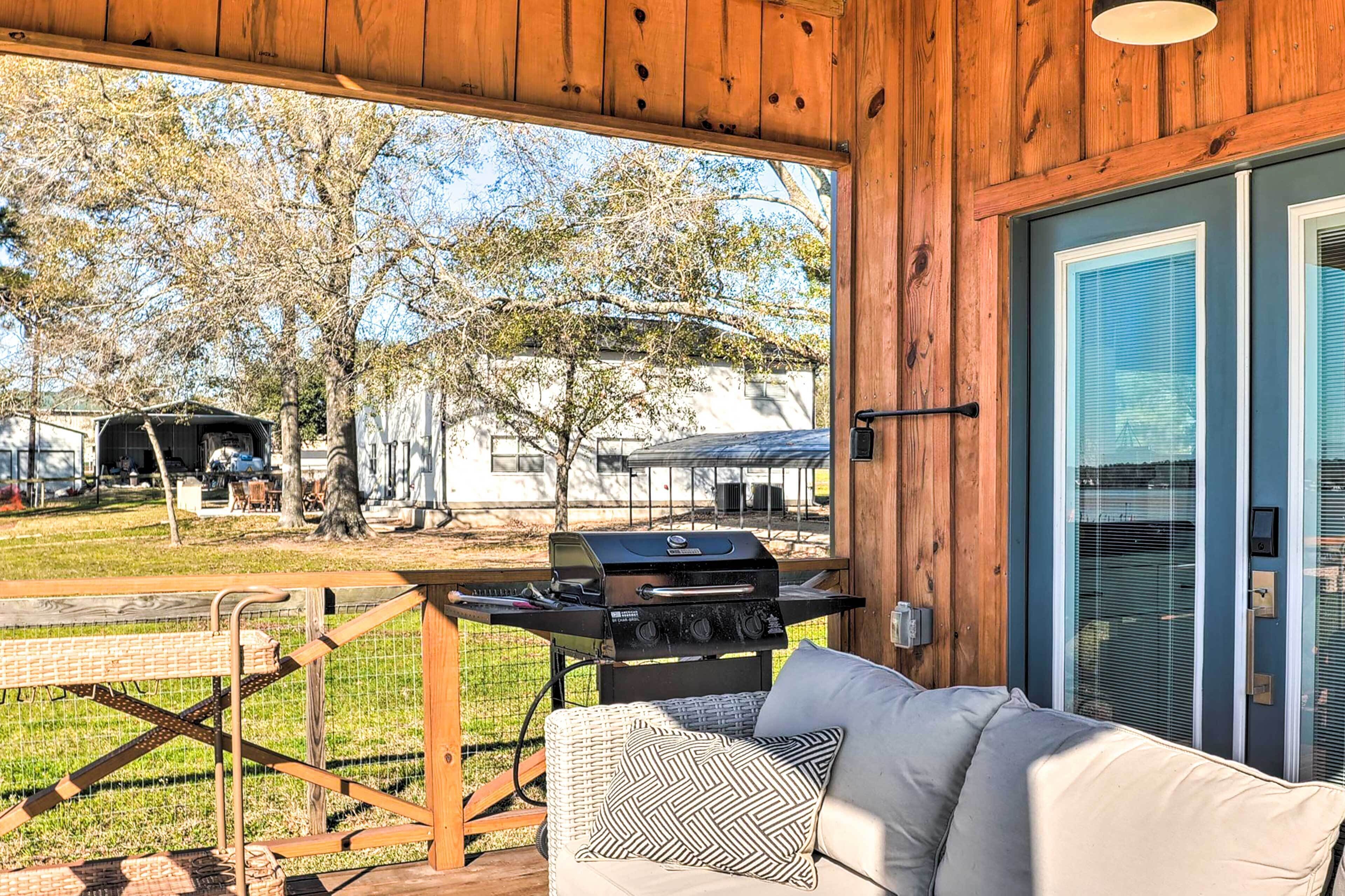 A covered outdoor area with a grill, a wicker sofa with cushions, and a view of a backyard with trees and a barbecue setup.