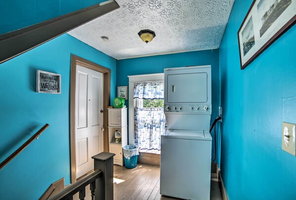 A laundry room with a white washing machine, a blue wall, a door, and a window with a curtain. - Explore Lake Erie & 'strip' From Quaint Apartment! (Geneva)