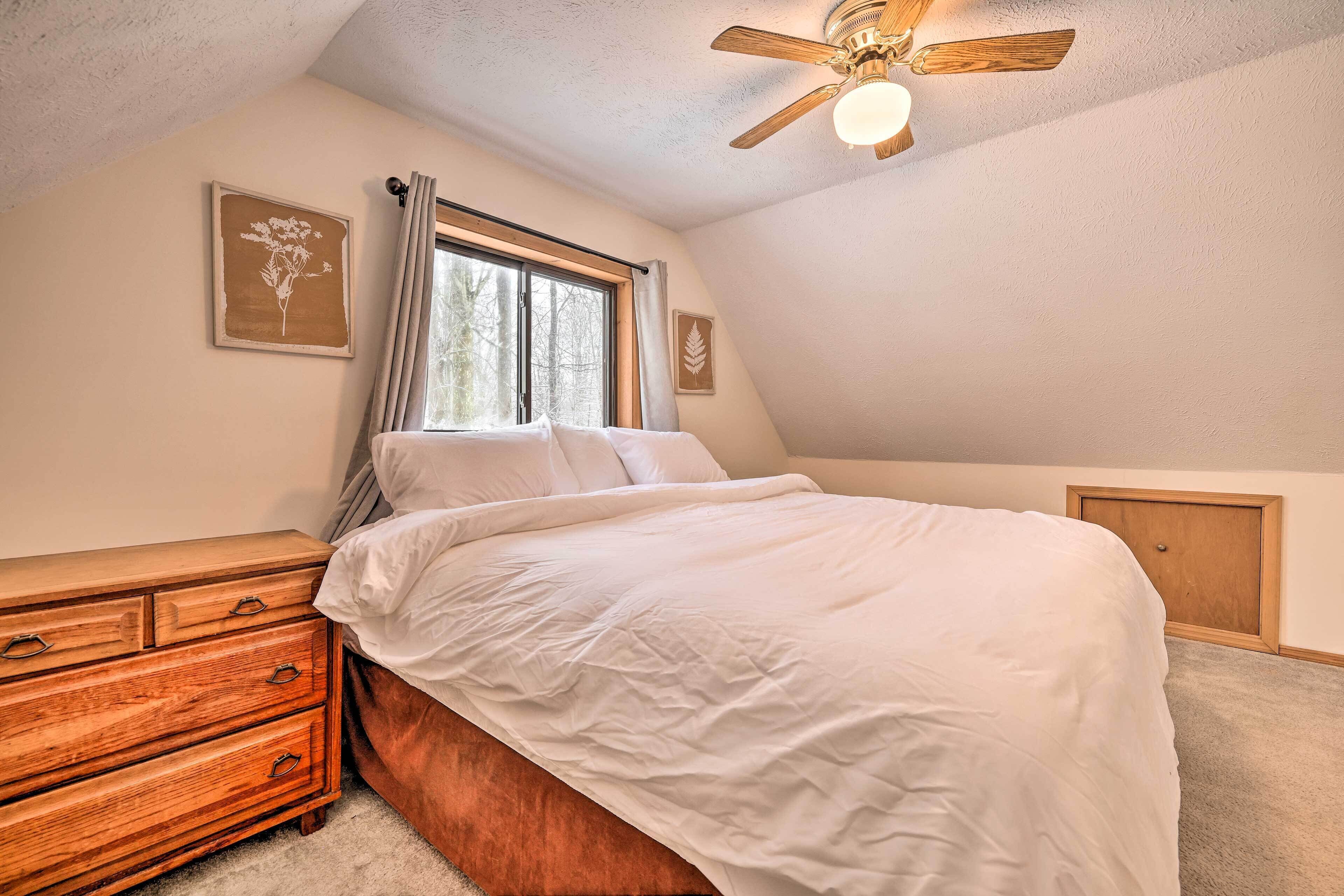 Une chambre à coucher avec un lit, une commode en bois, un ventilateur de plafond et une fenêtre avec des rideaux.