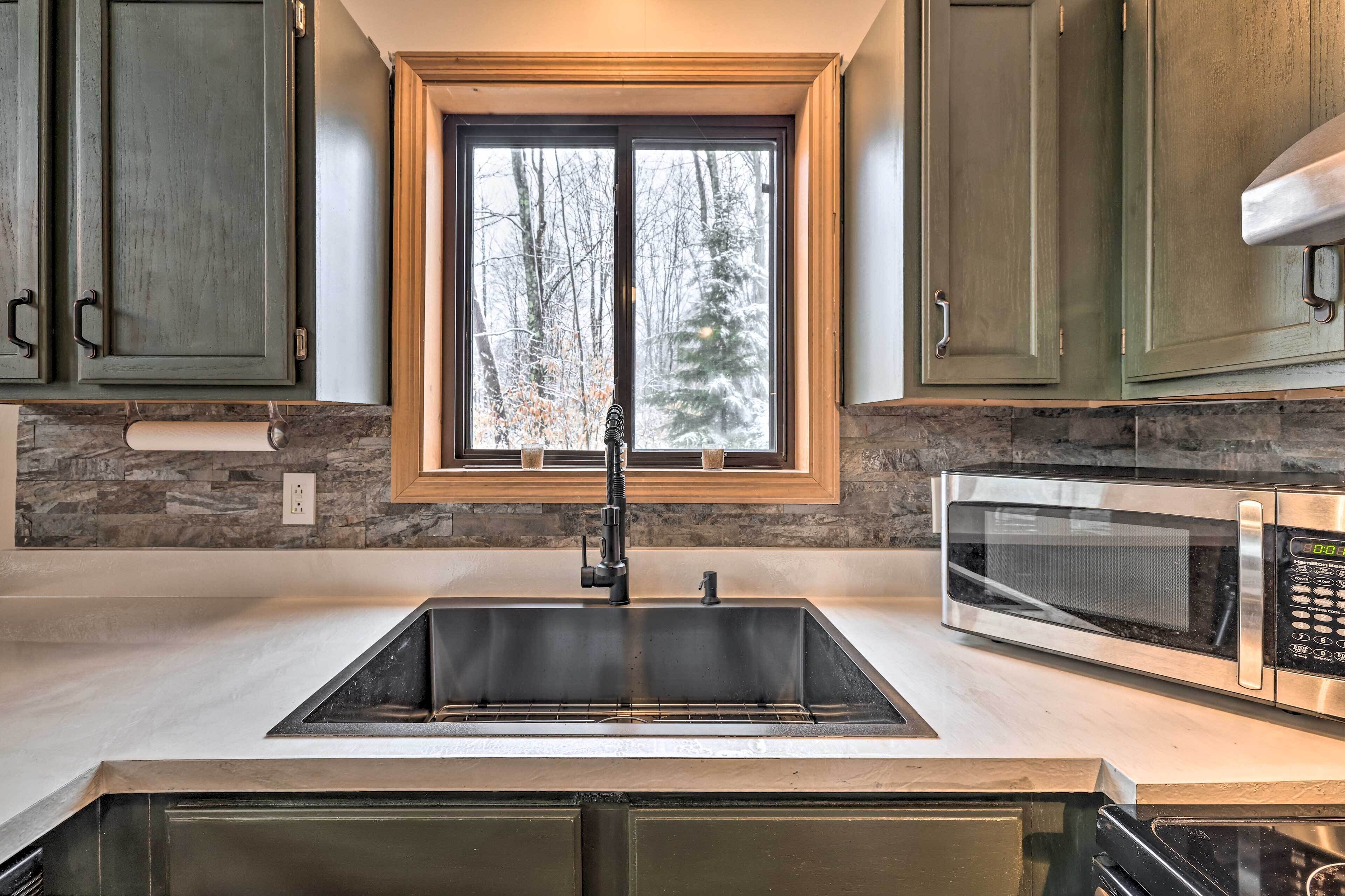 A kitchen with a double sink, a microwave, and a window with a view of trees.