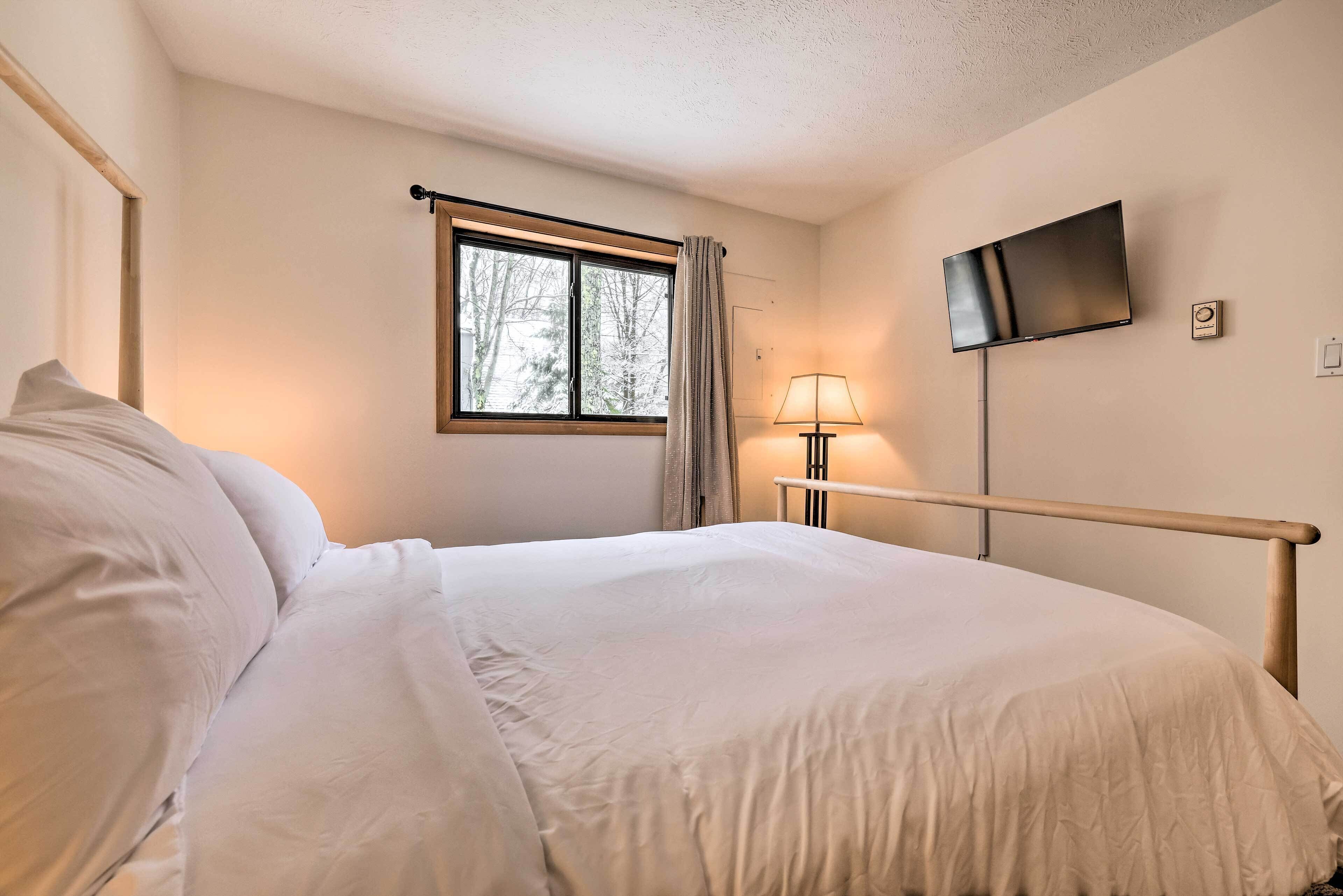 A neatly made bed with white linens, a bedside lamp, a flat-screen TV mounted on the wall, and a window with curtains.