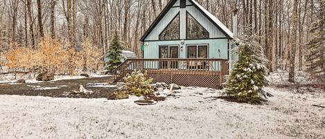 A cabin with a wooden deck and a large window, surrounded by a snowy landscape and trees.