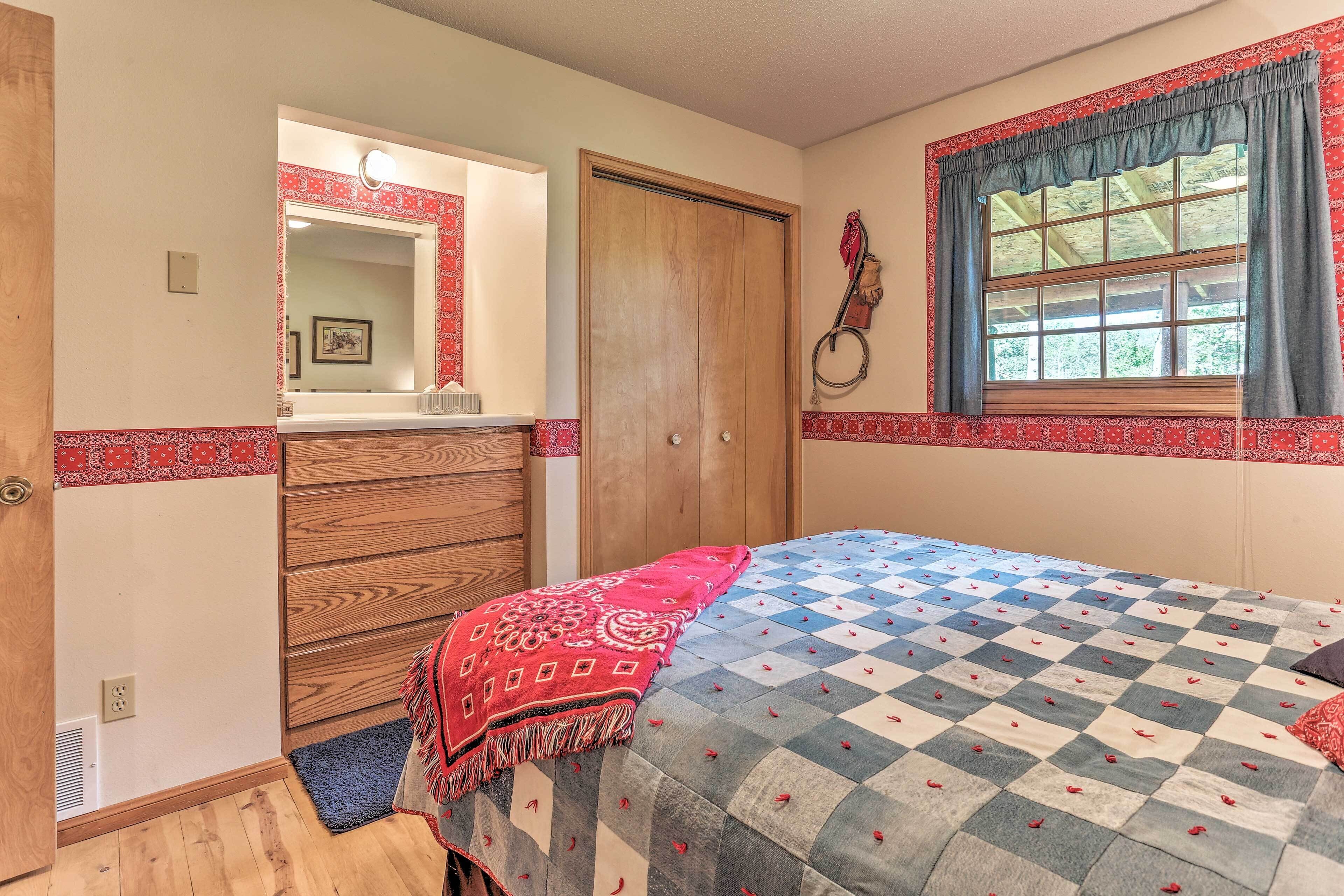 A bedroom with a wooden wardrobe, a bed with a patterned bedspread, a window with curtains, and a mirror with a red border.