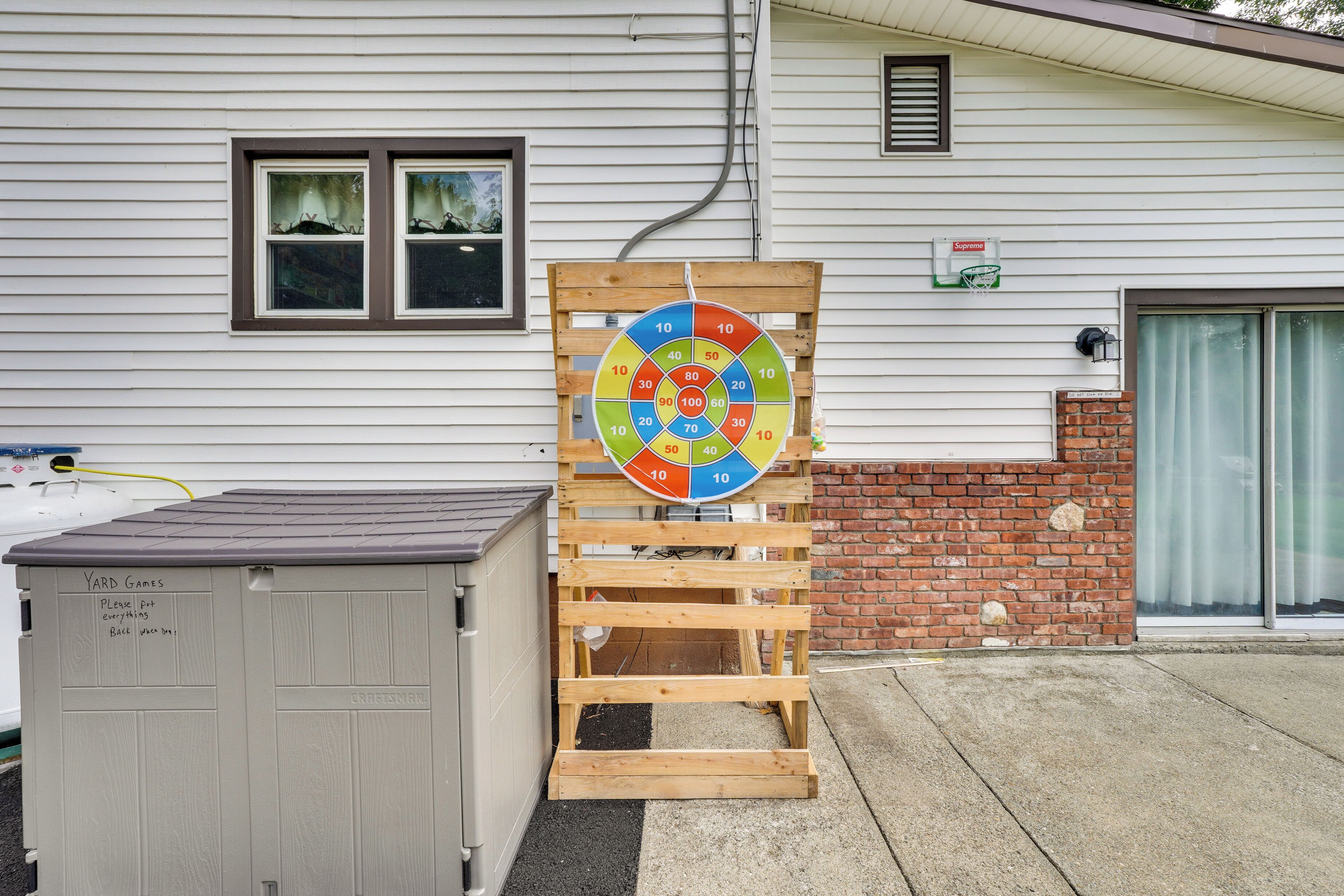 Een houten pallet met een kleurrijk dartbord, een tuinhuisje en een woning met een garage.