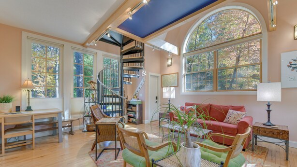 Un salon spacieux doté d’un plancher en bois, d’un canapé rouge, d’une table à manger avec des chaises, d’un escalier et de grandes fenêtres offrant une vue sur les arbres.