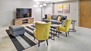 A modern living room with a sofa, two yellow chairs, a grey ottoman, a flat-screen TV, a wooden cabinet, and a floor lamp.