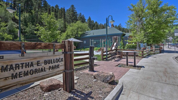 A wooden sign with "Martha E. Bullock Memorial Park" text, a paved walkway, and a playground area with a slide and benches.
