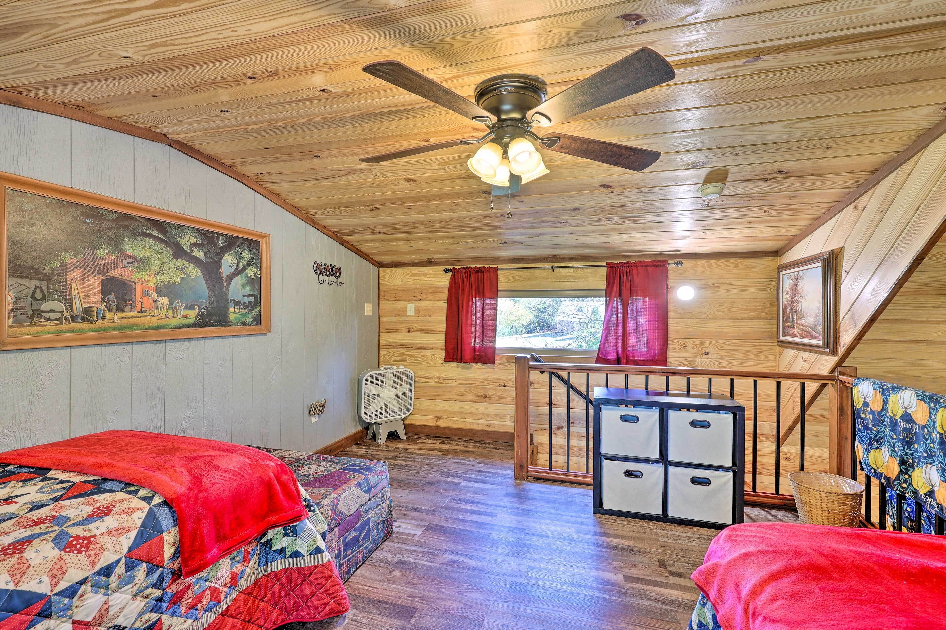 A cozy cabin room with a wooden ceiling, a ceiling fan, a bed with a red blanket, a small table, and a window with red curtains.