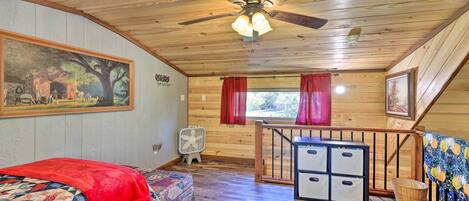 A cozy cabin room with a wooden ceiling, a ceiling fan, a bed with a red blanket, a small table, and a window with red curtains.