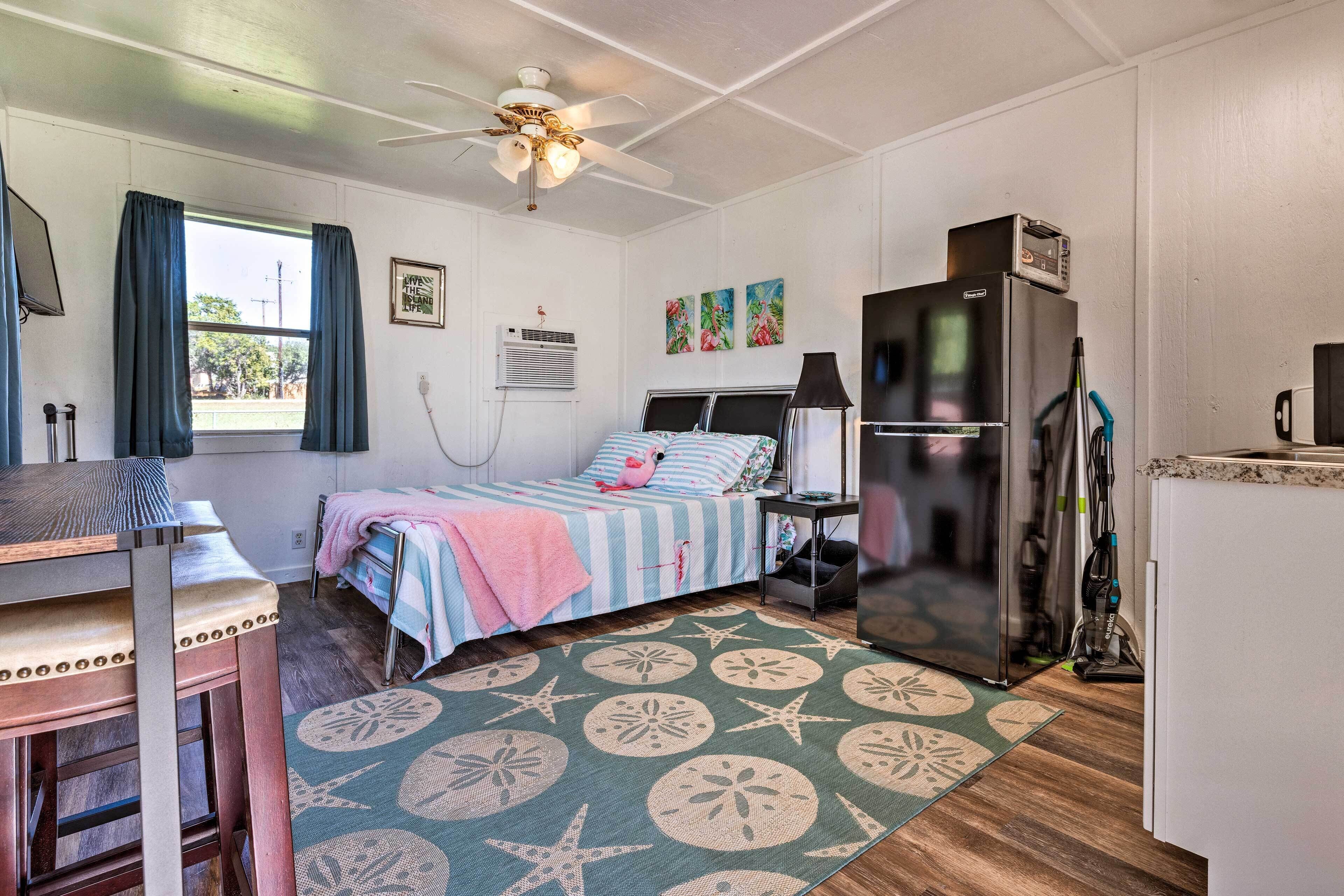 A compact room with a bed, a desk, a ceiling fan, a refrigerator, and a window with curtains.