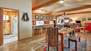 A spacious living area with a wooden dining table, chairs, and a red sofa. A kitchen with wooden cabinets and bar stools is visible in the background. - Dog-friendly Ranch Cottage w/ Yard ~ 13 Mi to Bend (Bend)
