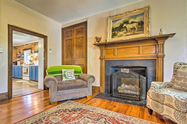 Una sala de estar con chimenea, dos sillones, un tapete estampado y una cocina al fondo.