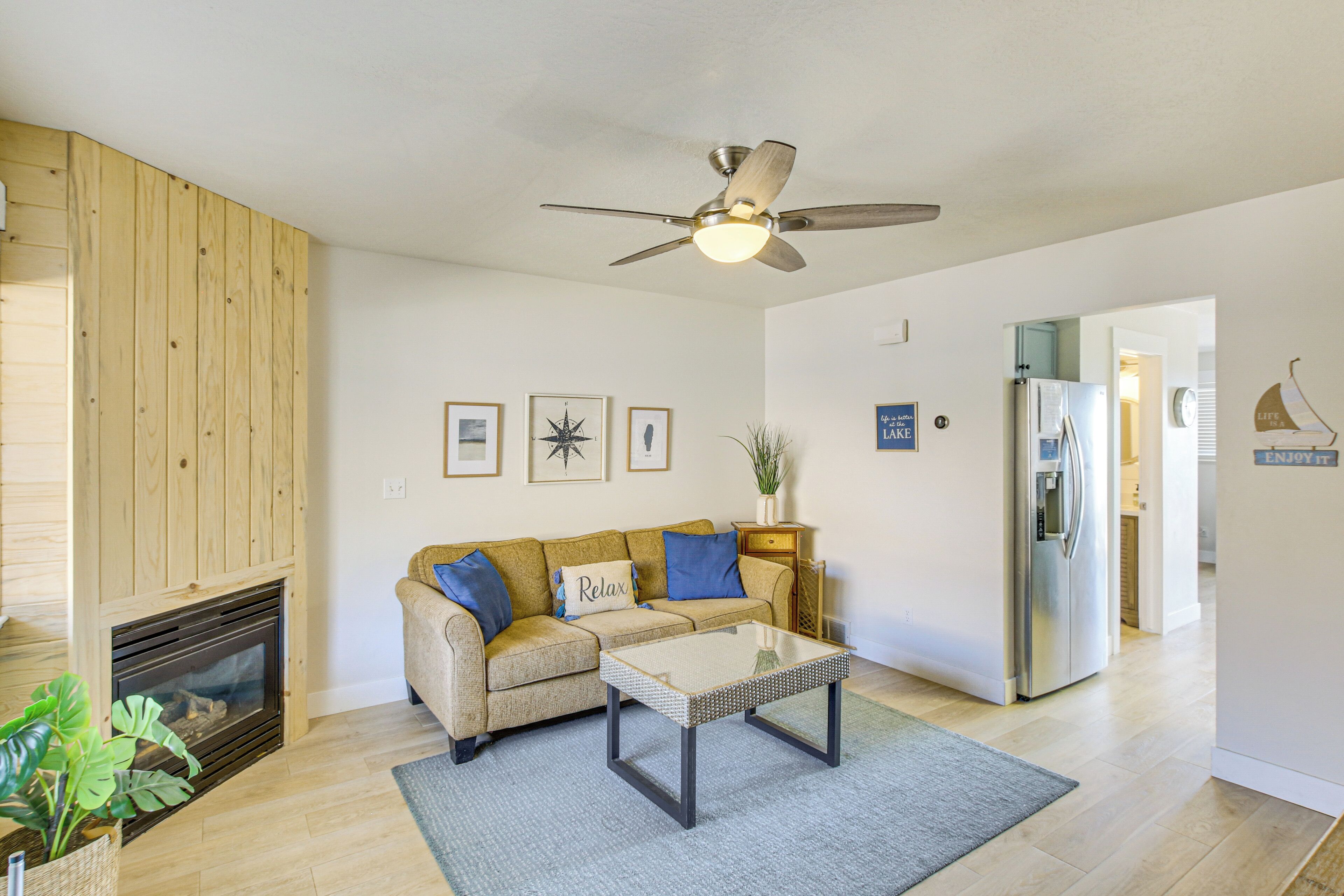 A living room with a beige sofa, a glass coffee table, a ceiling fan, a fireplace, and a refrigerator in the background.