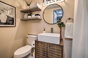 Un baño con un inodoro blanco, un mueble de madera con lavabo, un espejo y estantes con plantas y artículos de aseo.