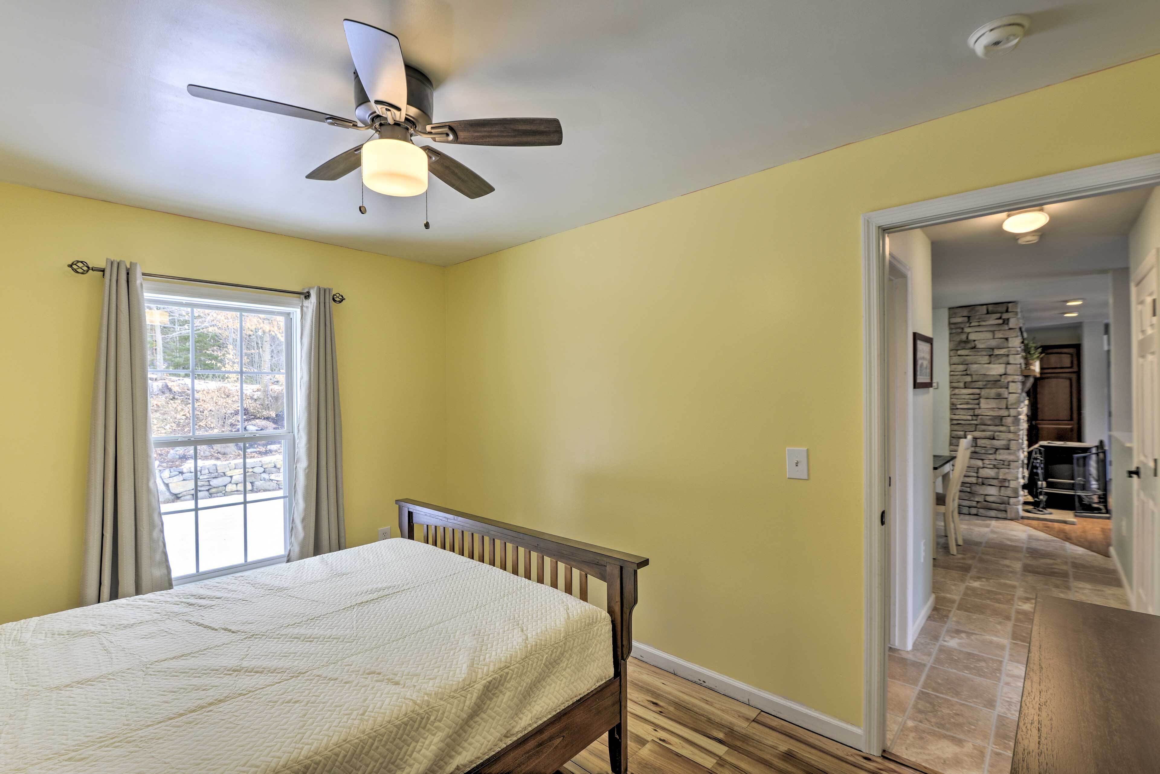 A bedroom with a bed, a ceiling fan, a window with curtains, and a view of a stone fireplace through an open door.