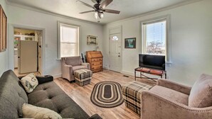 A living room with a ceiling fan, a sofa, a loveseat, a plaid ottoman, a television on a wooden stand, and a kitchen area visible through an open door.