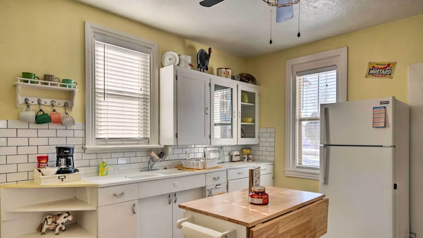 A kitchen with white cabinets, a wooden countertop, a refrigerator, and a window with blinds.