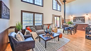 A spacious living area with a large couch, a coffee table, and a dining table set for two. There are large windows providing natural light and a view of the outdoors.