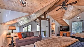 A spacious loft bedroom with a high ceiling, exposed wooden beams, a large bed, a television, and a cozy seating area with a sofa and a coffee table.
