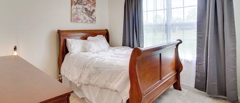 A bedroom with a wooden bed, a nightstand, a wall-mounted lamp, and a window with curtains.
