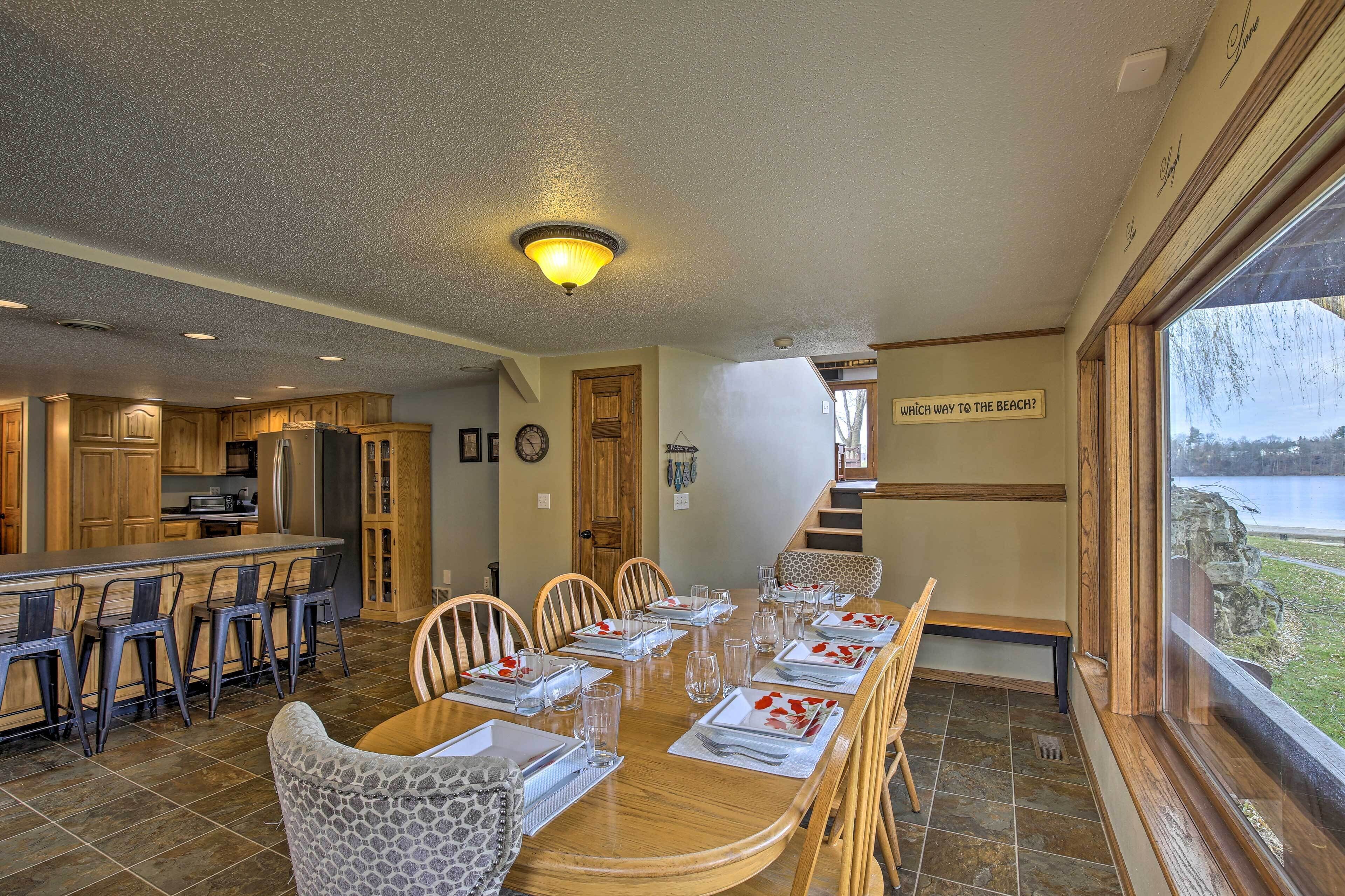 A dining area with a large table set for six, wooden chairs, and a view of a lake through a large window.