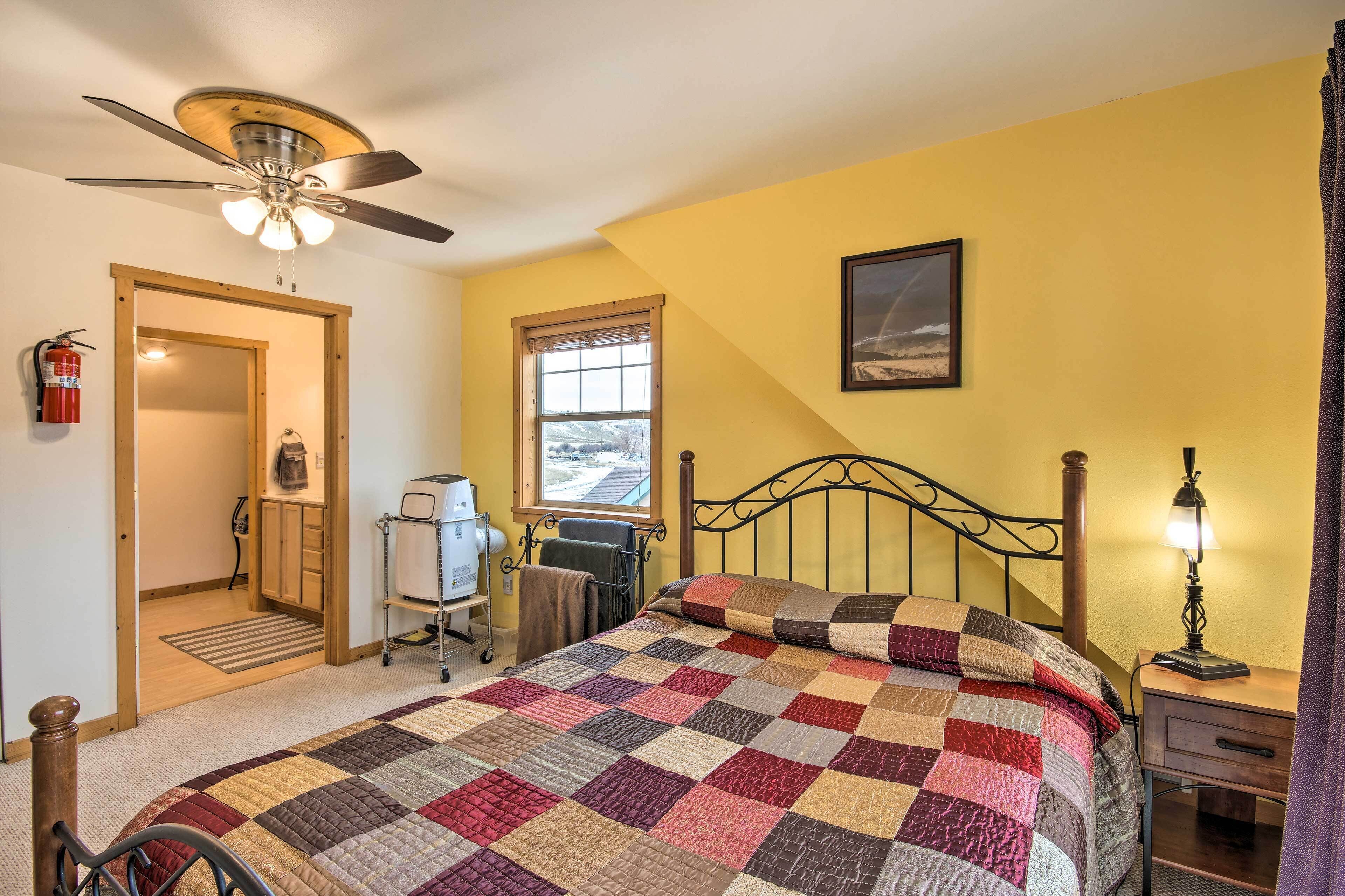 A bedroom with a bed, a ceiling fan, a window, a bedside table with a lamp, and a fire extinguisher on the wall.