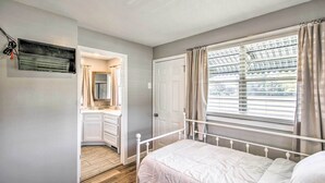 Een slaapkamer met een bed, een raam met gordijnen, een deur en een badkamer met een spiegel en wastafel.
