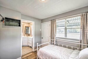 Een slaapkamer met een bed, een raam met gordijnen, een deur en een badkamer met een spiegel en wastafel.
