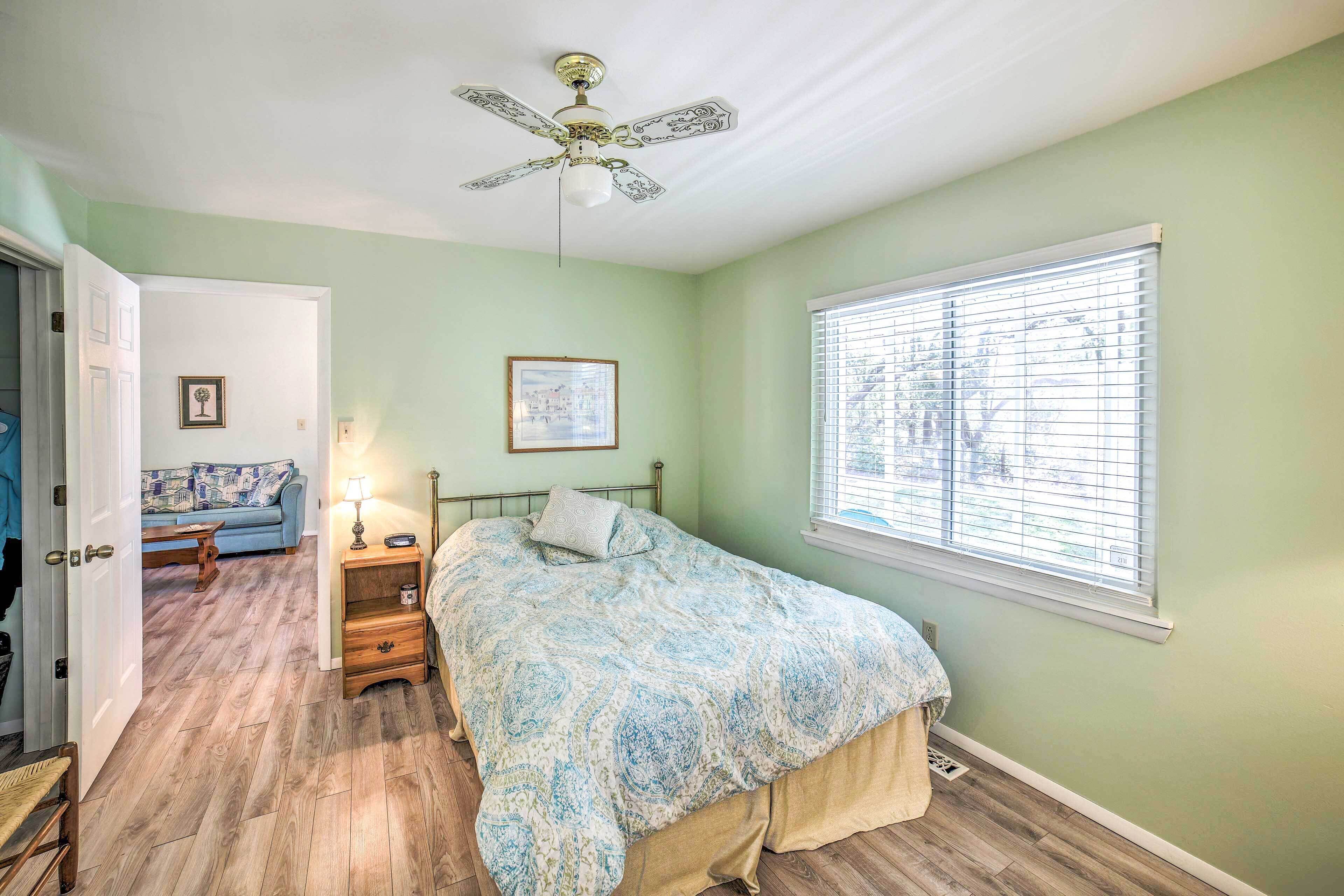 A bedroom with a bed, a ceiling fan, a window with blinds, a nightstand, a lamp, and a framed picture on the wall.