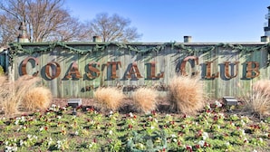 A wooden sign with "Coastal Club" written in large letters, surrounded by decorative plants and a clear blue sky. - Modern Lewes Home w/ Deck, Grill + Pond View! (Lewes)