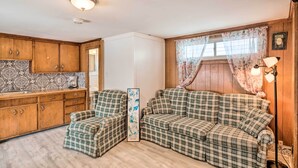 A cozy room with a plaid sofa, a matching armchair, and a small table. There is a window with floral curtains and a wall-mounted lamp. - No-frills Hampton Cottage - Steps to Beach! (Hampton)