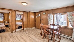 A room with wooden paneling, a wooden dining table with chairs, a bed with floral bedding, and a window with curtains. - No-frills Hampton Cottage - Steps to Beach! (Hampton)