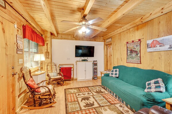 A cozy cabin interior with wooden walls, a ceiling fan, a television, a green sofa, rocking chairs, and a plaid rug.