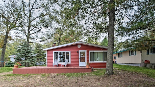 A small red cabin with a porch and two chairs, surrounded by trees and a grassy yard. - National City Cottage w/ Patio, Steps to Lake! (National City)