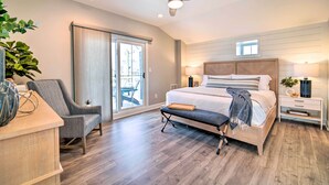 Une chambre à coucher comprenant un lit, des tables de chevet, une chaise, un banc, un ventilateur de plafond et une porte-fenêtre coulissante donnant sur un balcon.