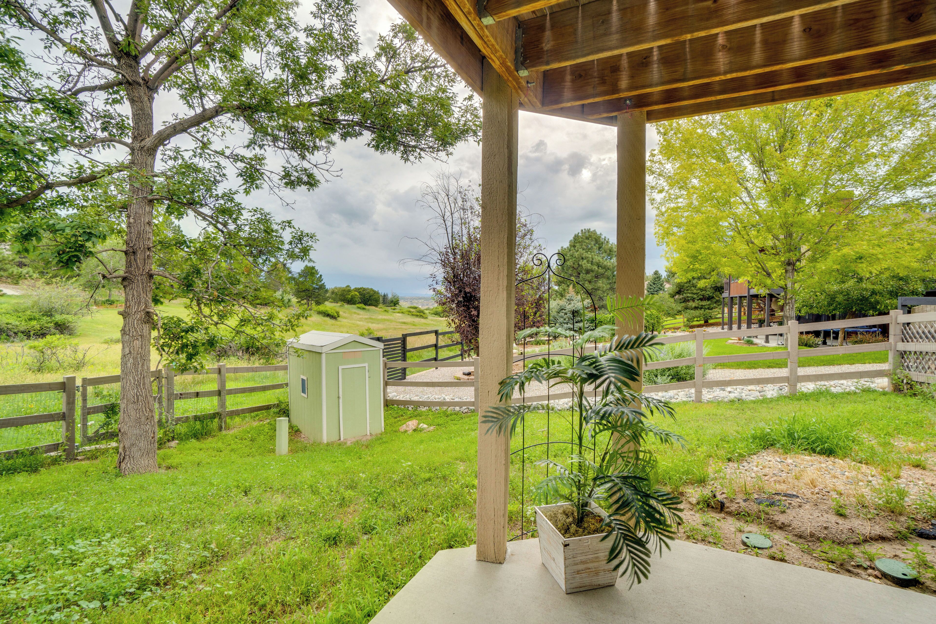 A covered patio with a view of a green landscape, a wooden fence, and a small shed.