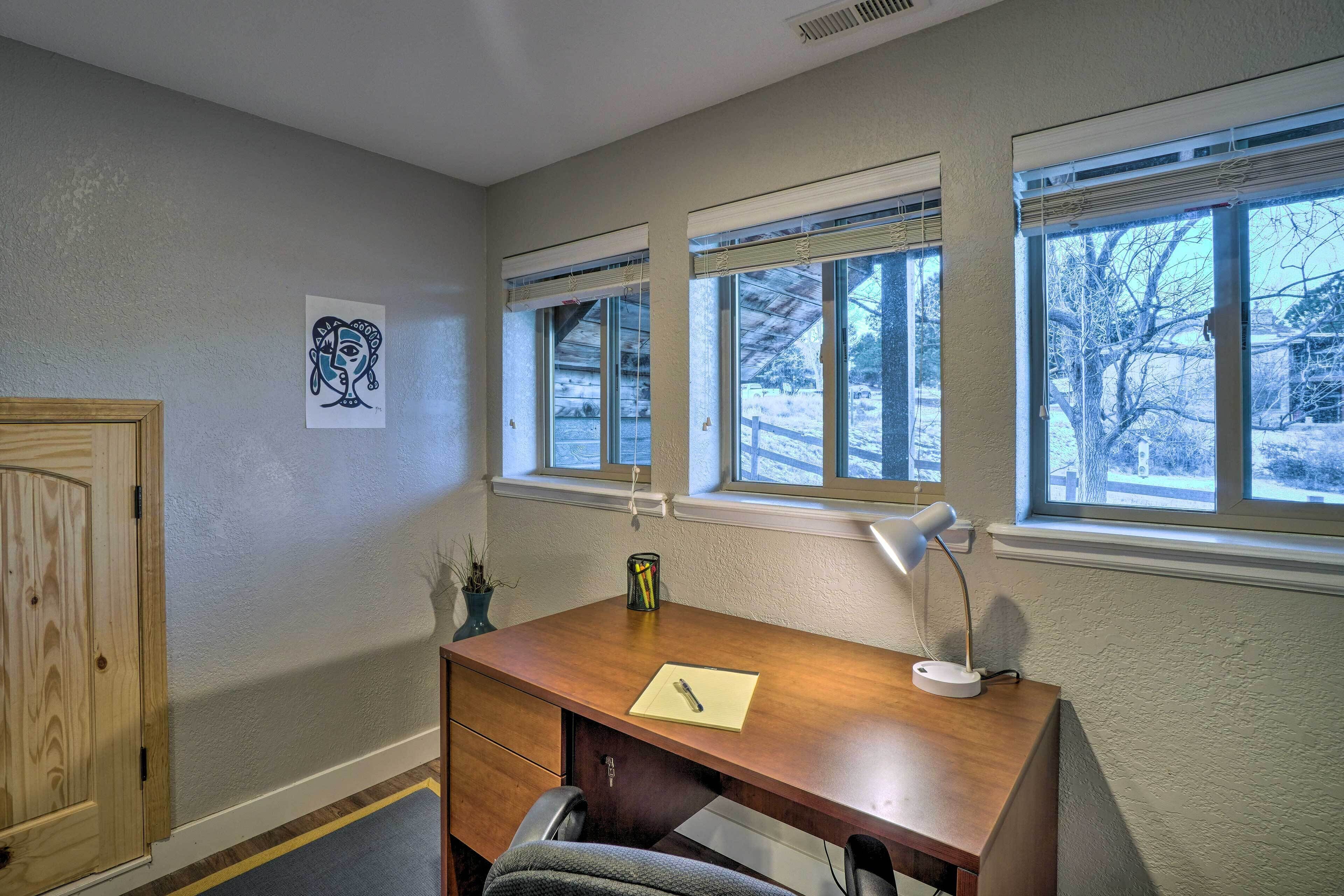 A home office with a wooden desk, a chair, a desk lamp, and a window with a view of trees.