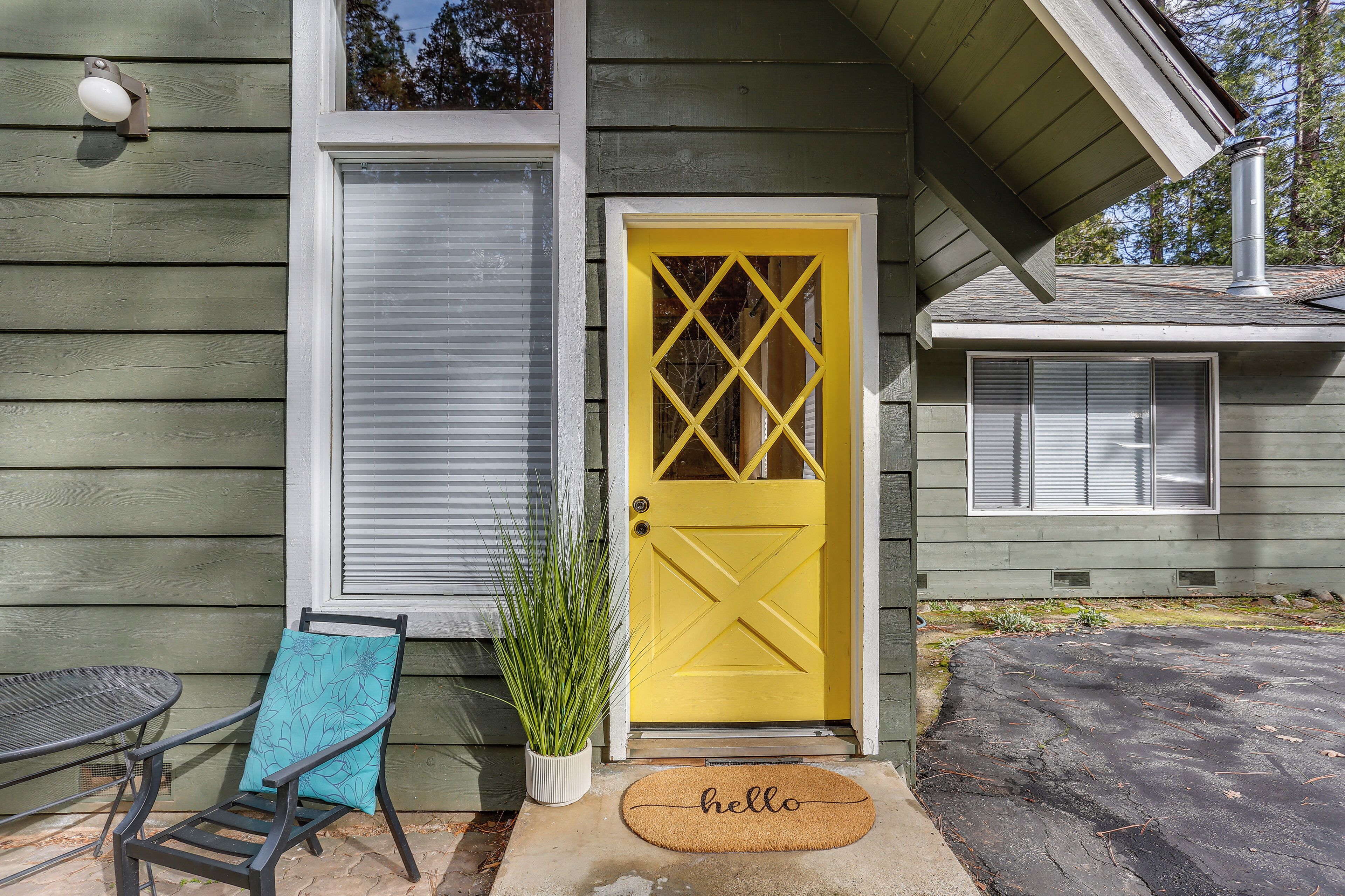 Une porte jaune avec une fenêtre au-dessus, un pot de fleurs, une chaise bleue et un paillasson sur lequel est écrit « bonjour », le tout sur un porche.