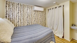 Una camera da letto con un letto, un cuscino bianco e soffice, una coperta a motivi, un condizionatore d'aria a parete, una tenda bianca e un piccolo ripiano con una radio.