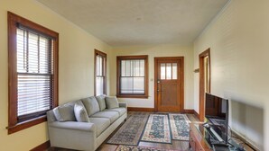 A living room with a sofa, two windows with blinds, a door, and a flat-screen TV. - Quaint Omaha Home < 5 Mi to Downtown! (Omaha)