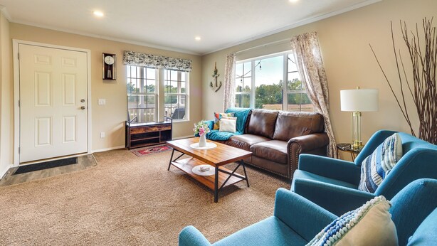 A living room with a brown leather sofa, a blue armchair, and a wooden coffee table. There is a window with curtains, a door, and a wall clock. - Family Home in Rapid City ~ 4 Mi to Torch Lake! (Rapid City)