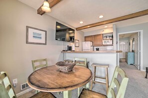 A dining area with a round wooden table, green chairs, and a basket. A kitchen is visible in the background with wooden cabinets and a microwave.