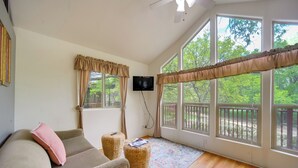 Una sala de estar acogedora con un sofá beige, un cojín rosa, un televisor de pantalla plana, un ventilador de techo y ventanas amplias con vistas a zonas verdes.