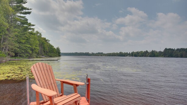 A wooden dock with a single Adirondack chair facing a calm lake, surrounded by dense forest. - Rhinelander Home - Jennie Webber Lake On-site (Rhinelander)