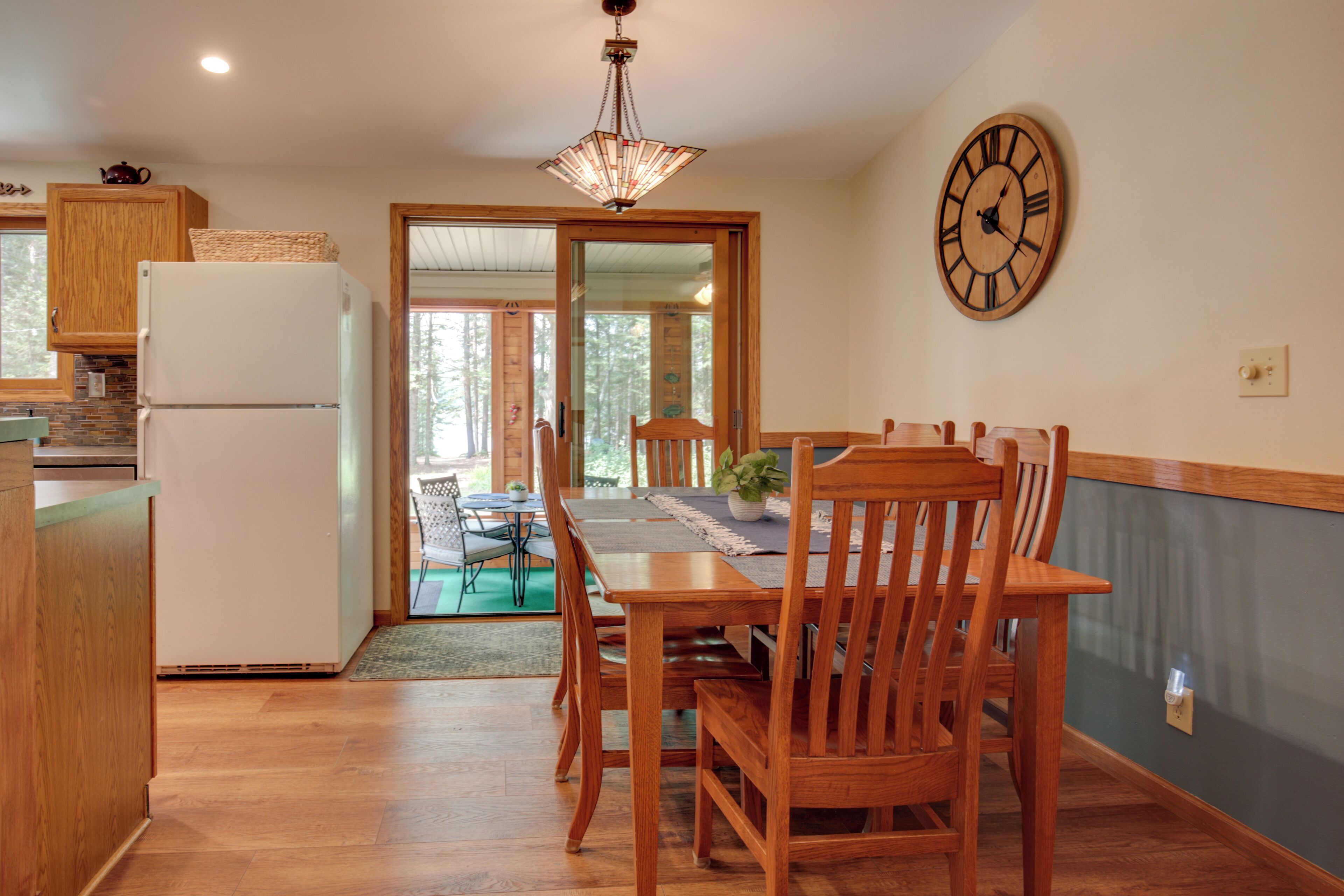 Een keuken met houten kastjes, een witte koelkast en een eettafel met stoelen. Er hangt een klok aan de muur en een deur leidt naar een balkon.