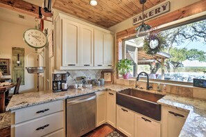 A kitchen with white cabinets, a stainless steel dishwasher, a coffee maker, and a window with a view of trees. - Secluded Country Hideaway w/ Fire Pit & Pool! (Blanco)