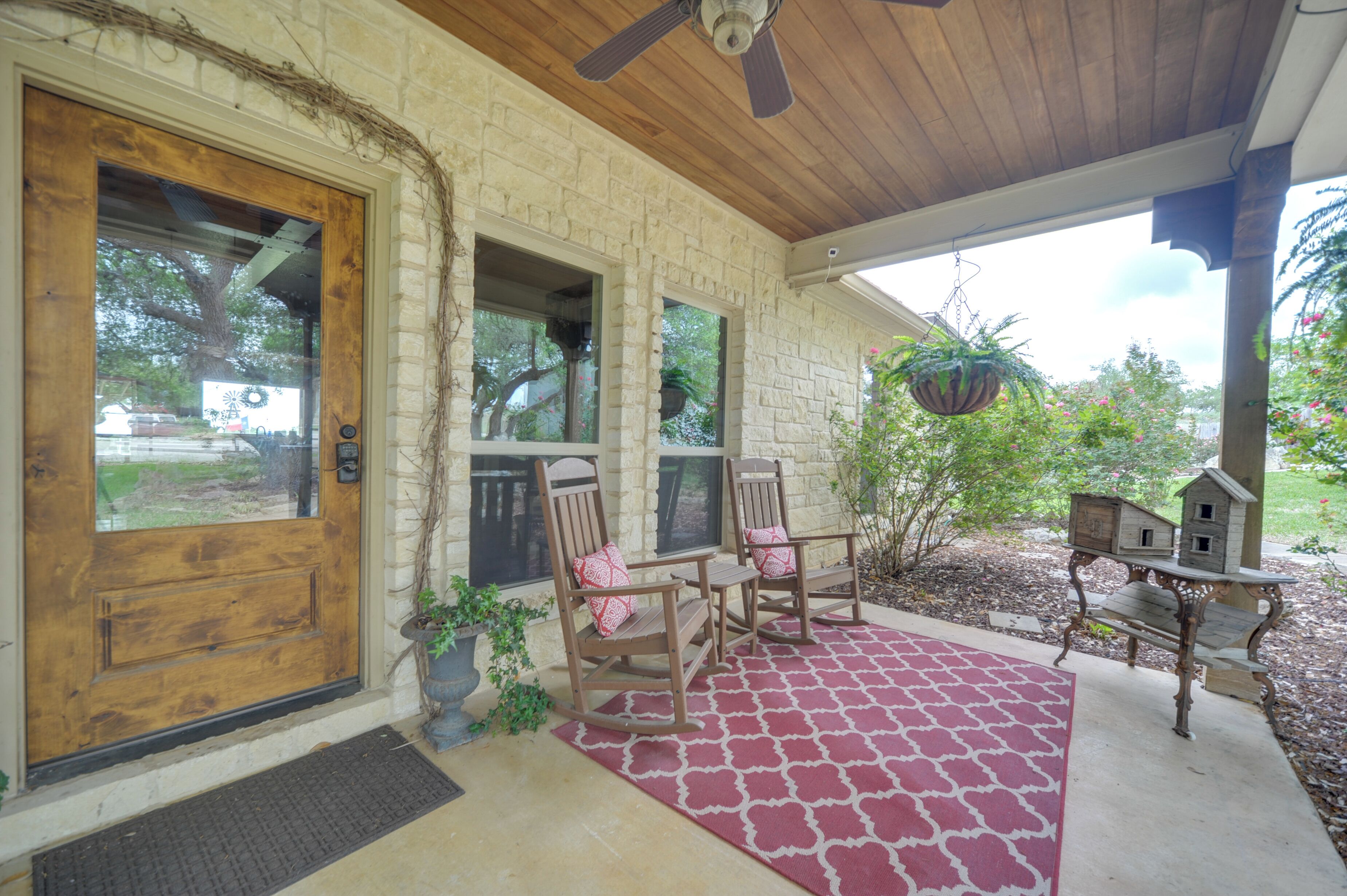 A stone house with a wooden door, a porch with rocking chairs, a red patterned rug, and a birdhouse on the right.