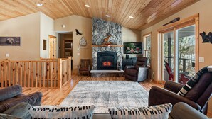 A cozy living room with a fireplace, wooden ceiling, and a view through large windows.