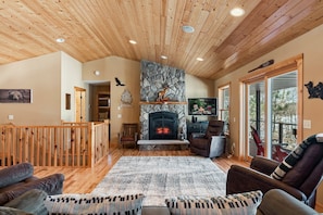 A cozy living room with a fireplace, wooden ceiling, and a view through large windows. - Lake Ossie| Kayaks| Great Swimming | Firepit (Breezy Point)
