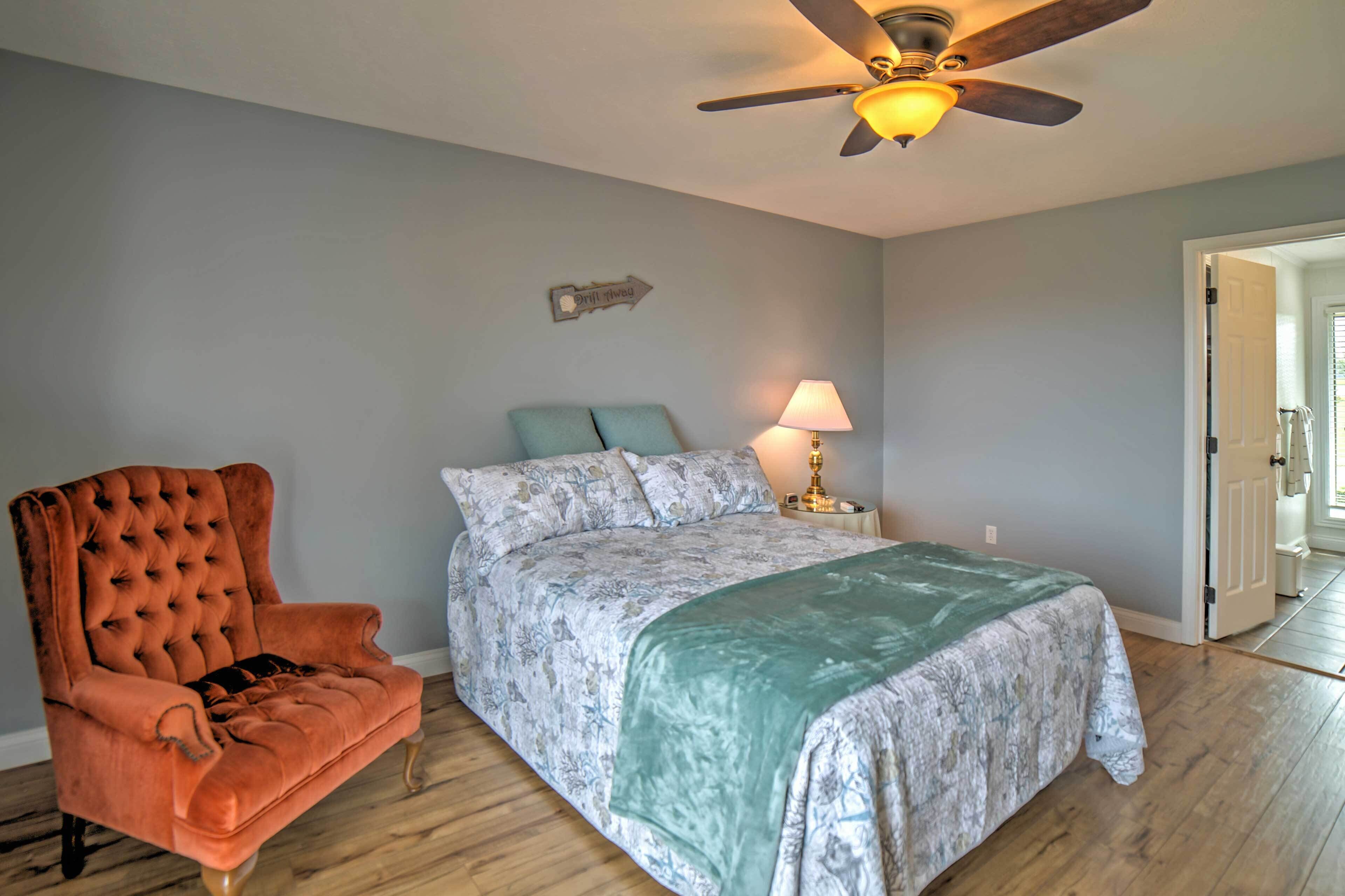 A bedroom with a tufted armchair, a bed with a patterned bedspread, a nightstand with a lamp, and a ceiling fan.